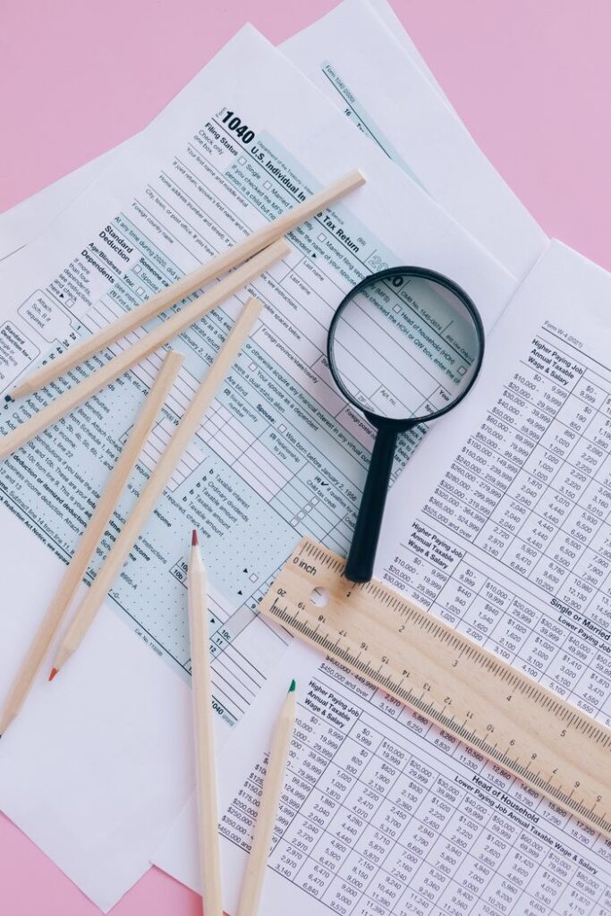 Tax documents with magnifying glass and pencils on a pink surface.